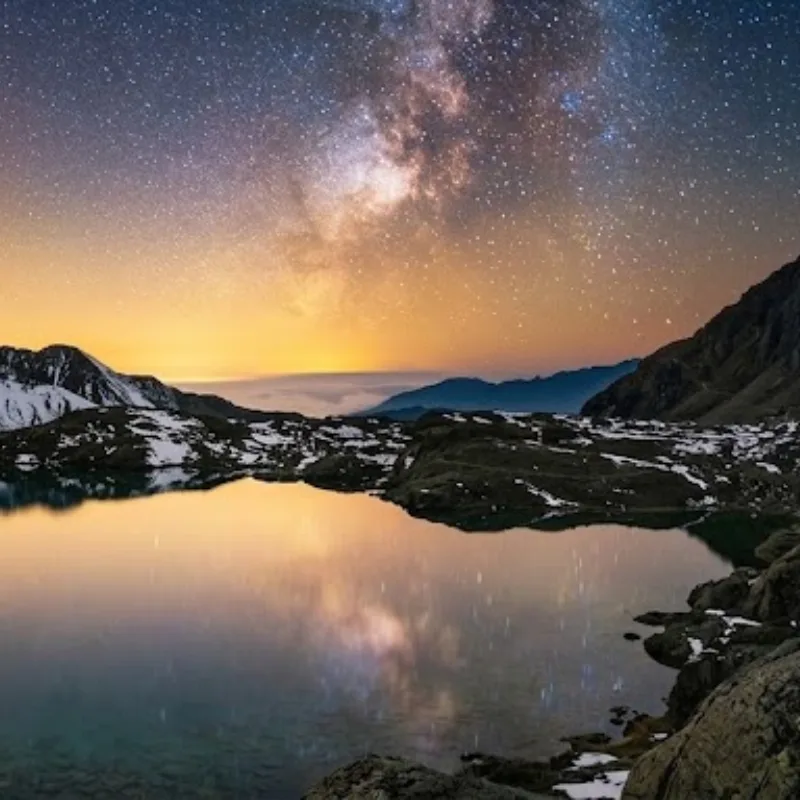 Night sky with the Milky Way and golden horizon reflected in a calm high-altitude alpine lake surrounded by snow-dusted mountains.