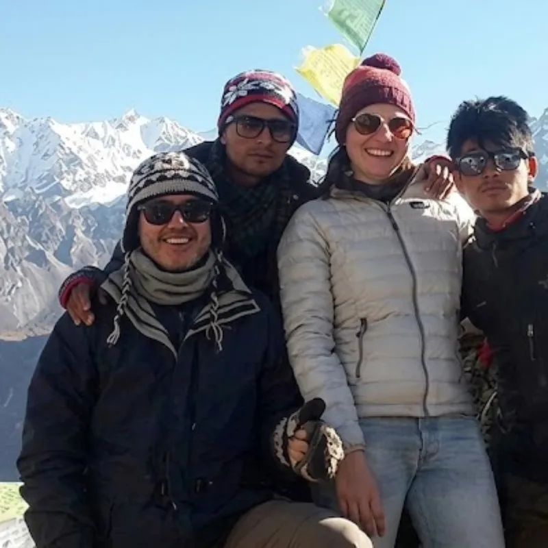 Group of four trekkers wearing beanies and sunglasses smiling with colorful Buddhist prayer flags and snowy peaks behind them.