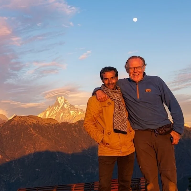 Two smiling trekkers standing outdoors against the sunlit Himalayan peak of Machapuchare under a blue morning sky.