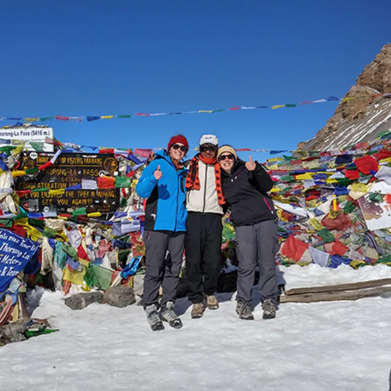 Three smiling trekkers posing in the snow in front of the Thorong-La Pass elevation sign at 5416 meters, heavily draped in colorful Buddhist prayer flags