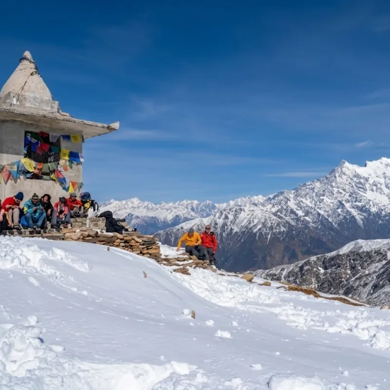 Trekkers resting near a traditional Buddhist stupa covered in snow with Himalayan peaks in the background.