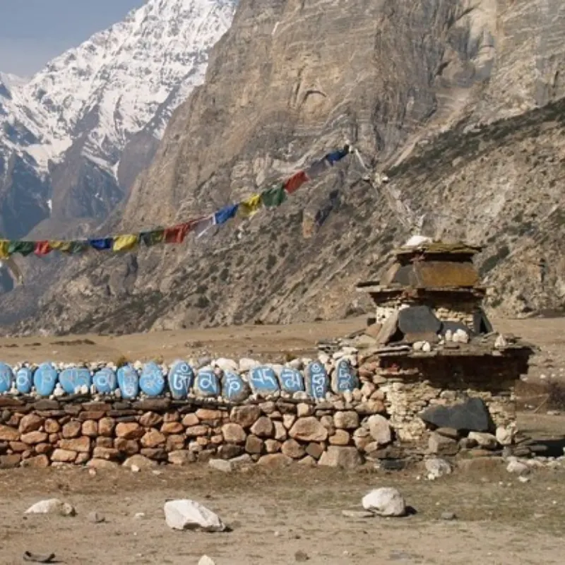 A traditional stone Mani wall featuring blue stones carved with Tibetan mantras, alongside a stone chorten and colorful prayer flags in a rugged mountain landscape.