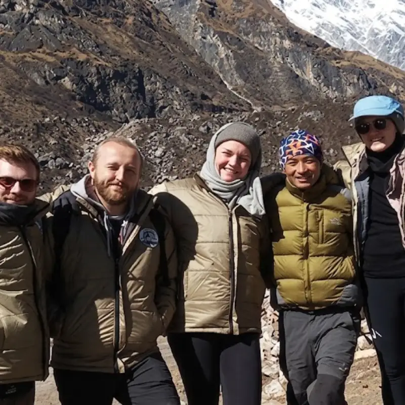 A diverse group of five smiling trekkers standing outdoors in warm down jackets against a rocky mountain terrain.