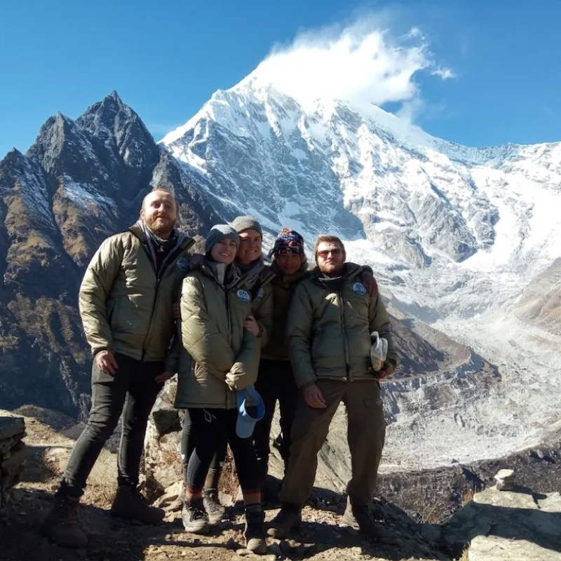 A group of male and female hikers wearing heavy winter jackets smiling together on a rocky ridge with the massive snow-covered peak of Annapurna rising behind them.