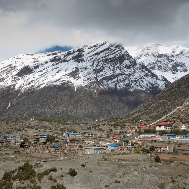 Traditional stone village nestled beneath dramatic snow-dusted rock faces and dark clouds in the high Himalayas Nepal.