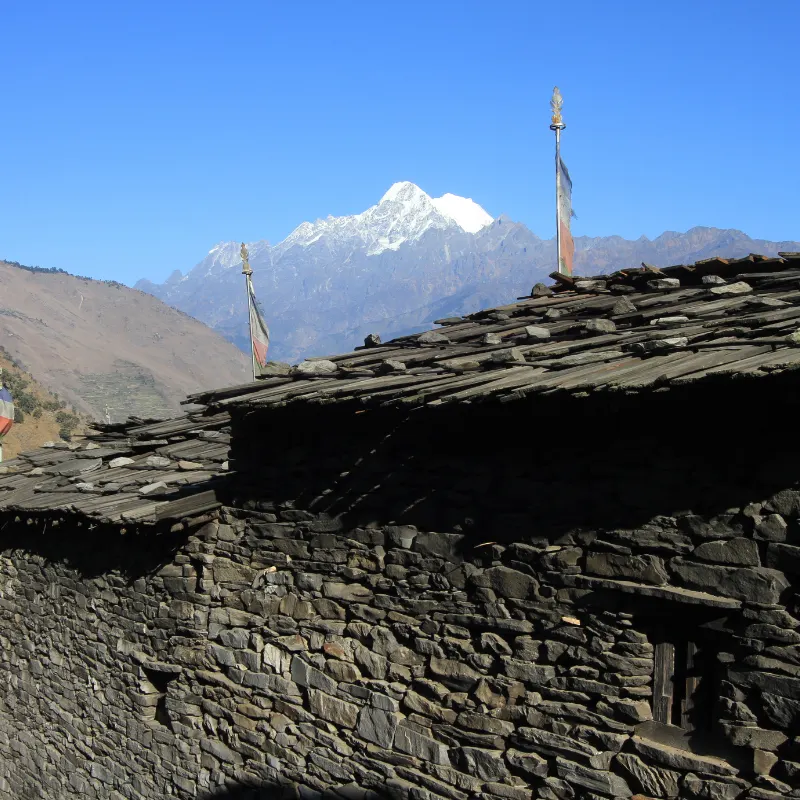 Traditional Himalayan rustic stone house with a slate roof and prayer flags overlooking distant snow-capped mountain peaks.