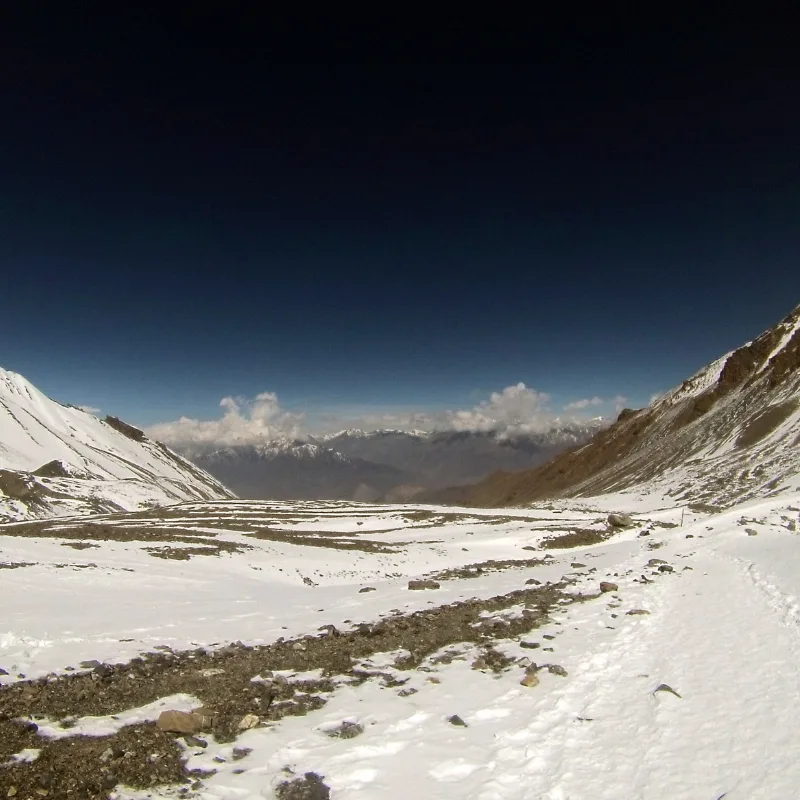 A snow-covered trekking trail cutting through a wide mountain pass, looking down toward distant arid valleys beneath a deep blue, high-altitude sky.