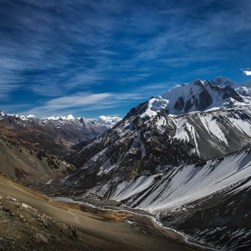 A rugged, snow-dusted alpine valley descending toward a backdrop of massive, snow-capped Himalayan peaks beneath a bright blue sky.