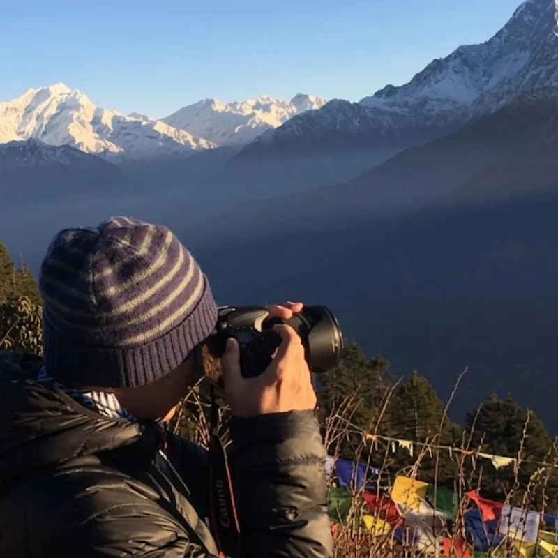 Photographer in a knit beanie aiming a DSLR camera toward the snow-capped Himalayan mountain ridges at sunrise.