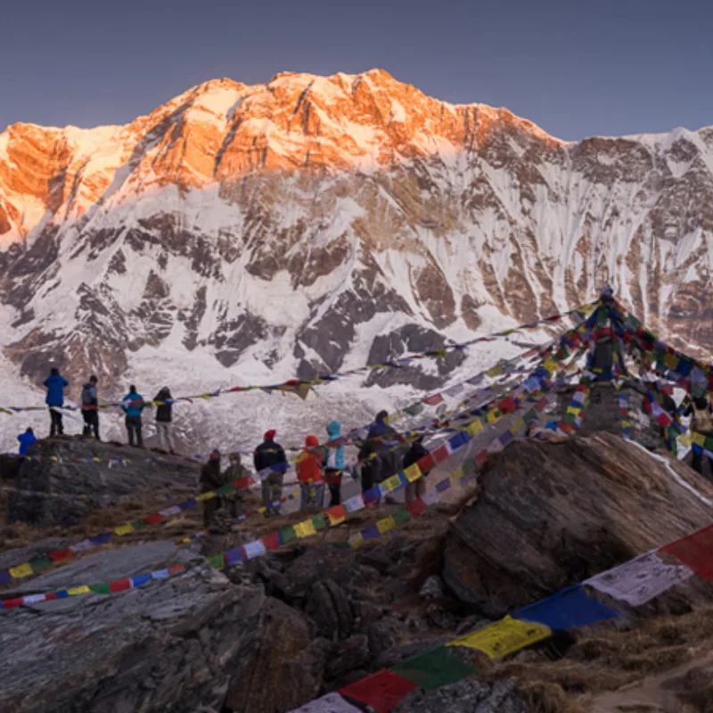 Trekkers gathered at Annapurna Base Camp watching the golden morning light hit the south face of Annapurna with prayer flags in the foreground.
