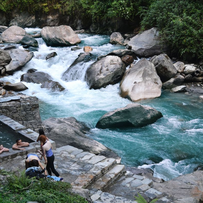 Trekkers relaxing in riverside natural hot springs next to turquoise glacial river rapids in Jhinu Danda Nepal.