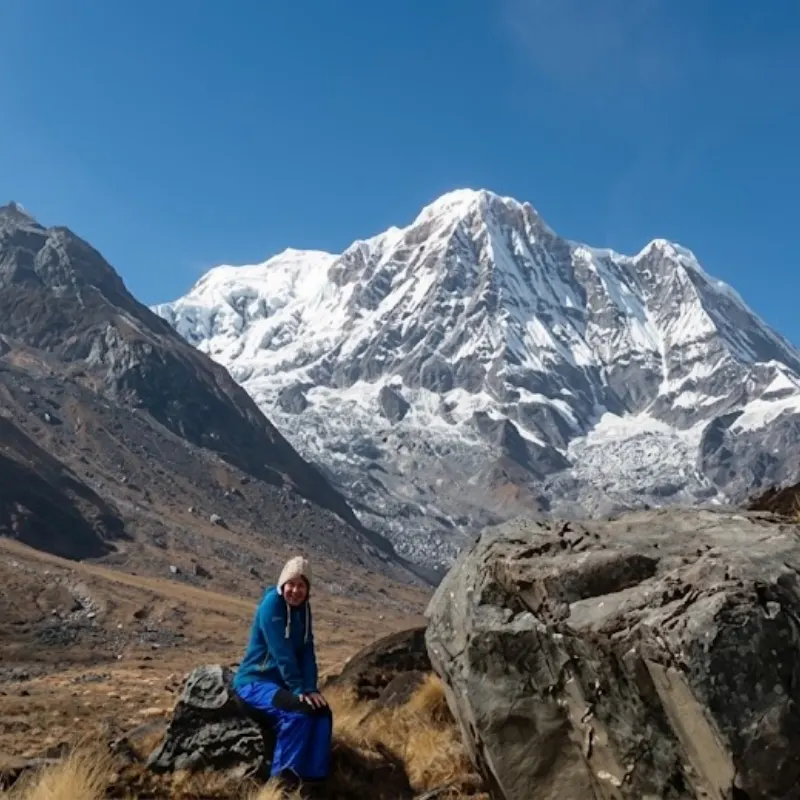 A trekker resting on a large rock in an open alpine valley with the snow-covered peak of Annapurna South rising in the background.