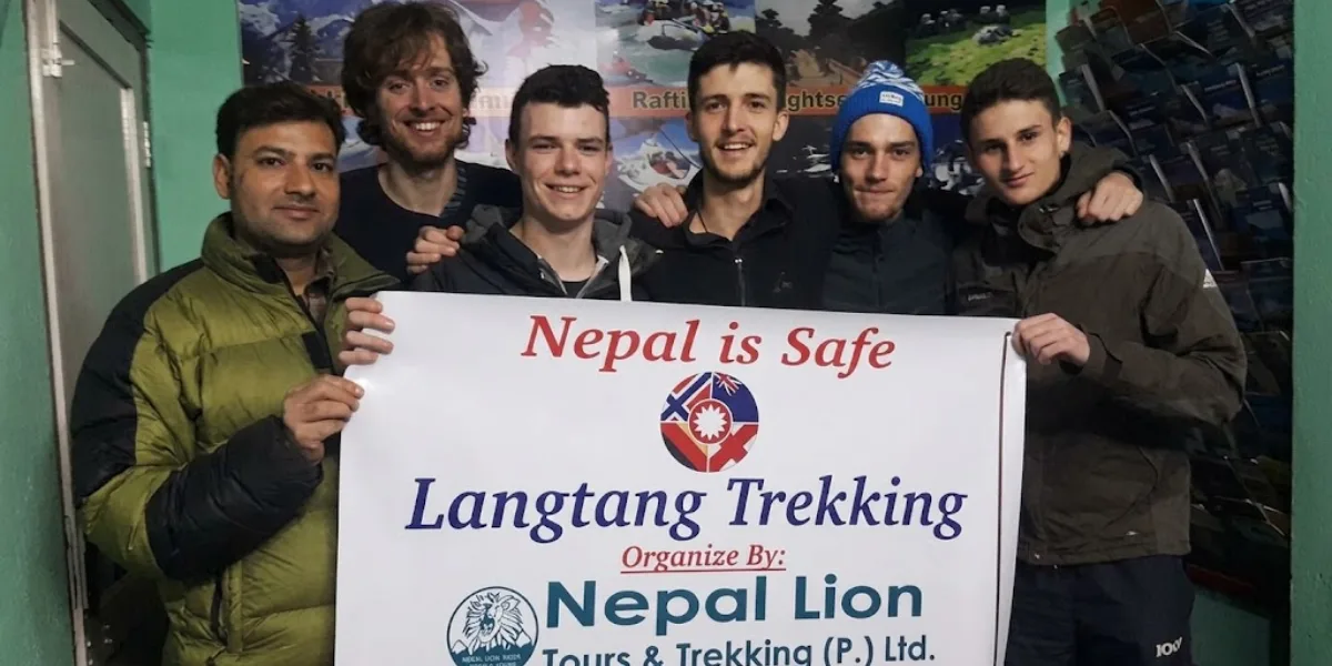 A group of smiling young male trekkers indoors holding a white banner reading "Nepal is Safe - Langtang Trekking Organized By Nepal Lion Tours and Trekking.