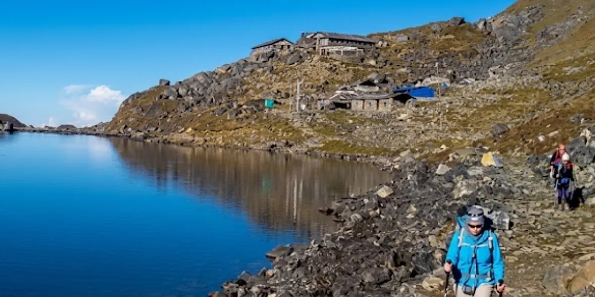 Trekkers walking along a rocky path next to a clear blue alpine lake with stone mountain lodges on the hillside.