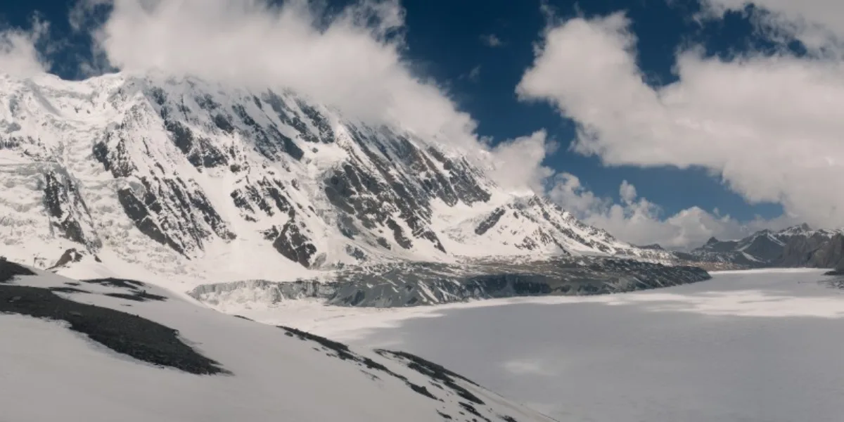A vast, flat frozen glacial plain sitting at the base of an imposing, heavily snow-covered mountain face draped in low-hanging clouds.