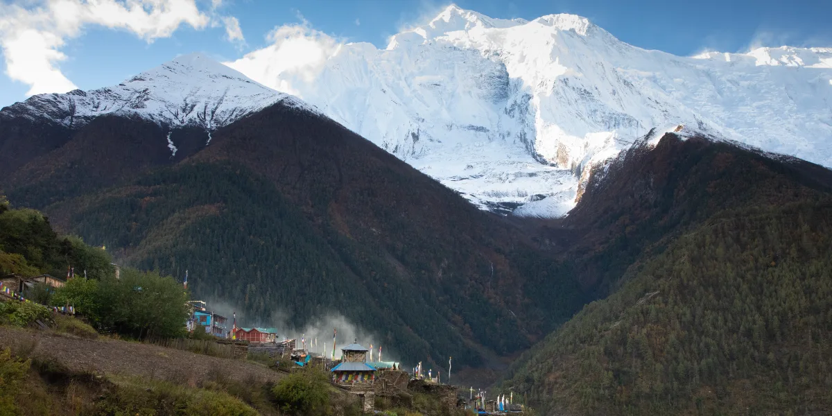 Traditional mountain village with colorful roofs and prayer flags sitting in a forested valley beneath a towering snow-covered Himalayan peak in Nepal.